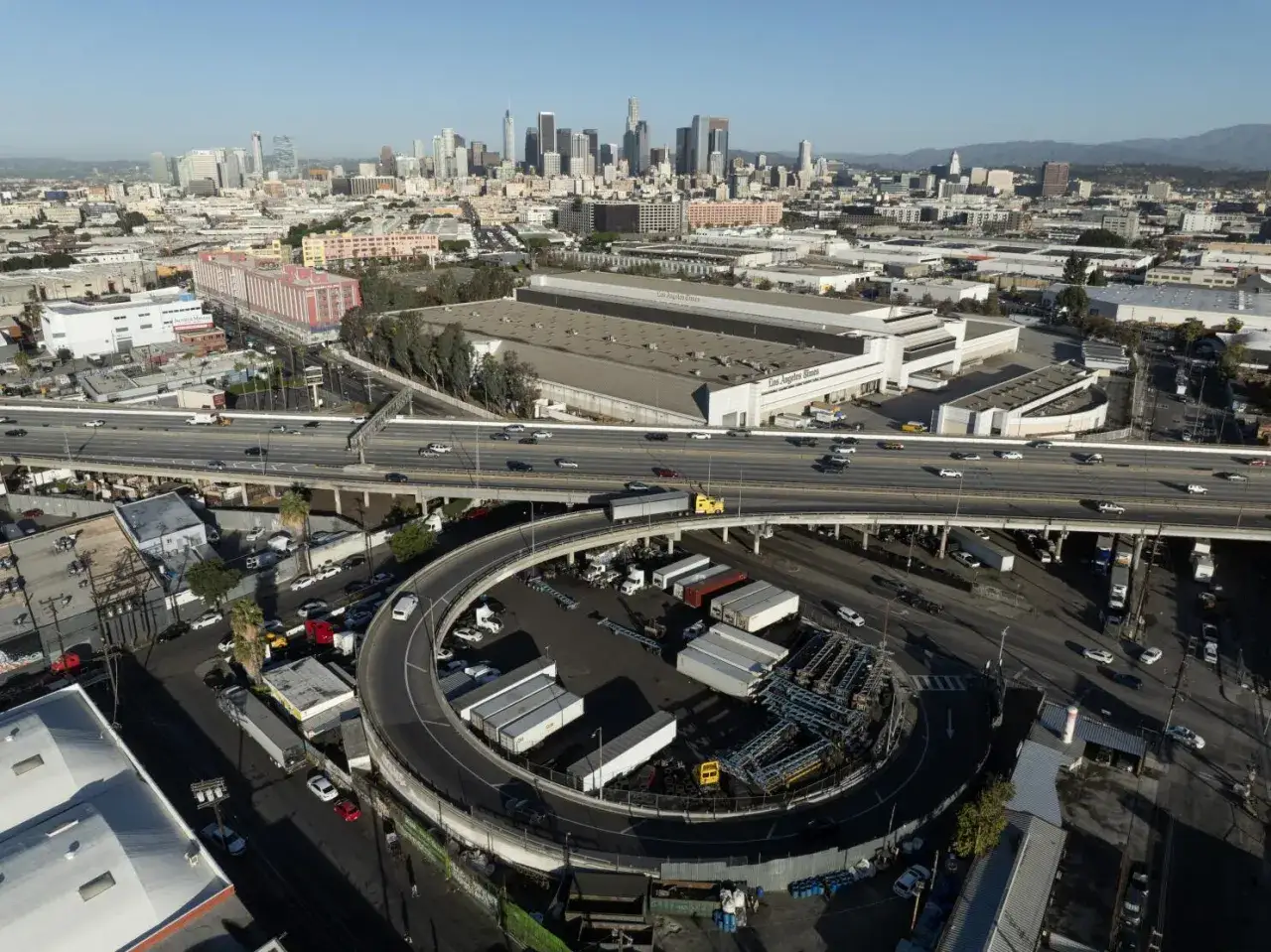 Vue a&eacute;rienne d'une autoroute &agrave; Los Angeles, avec des voitures circulant et un &eacute;changeur en forme de spirale. La skyline de la ville se dresse &agrave; l'horizon.