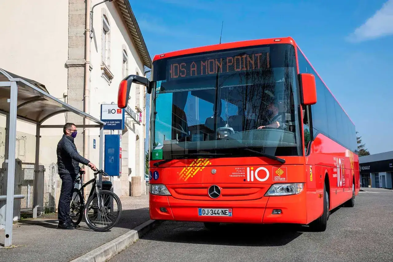 Un homme avec un vélo attend un bus rouge LIO à une station. Ce bus dessert les lignes Graulhet Gaillac.
