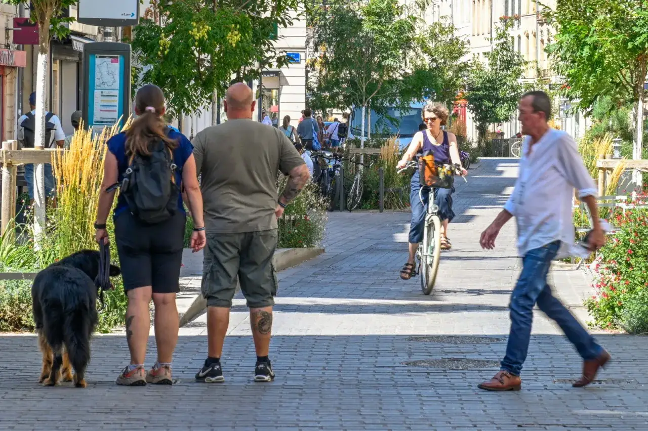 Un homme à vélo, un couple avec un chien et un homme qui marchent illustrent les mobilités actives dans une rue animée.