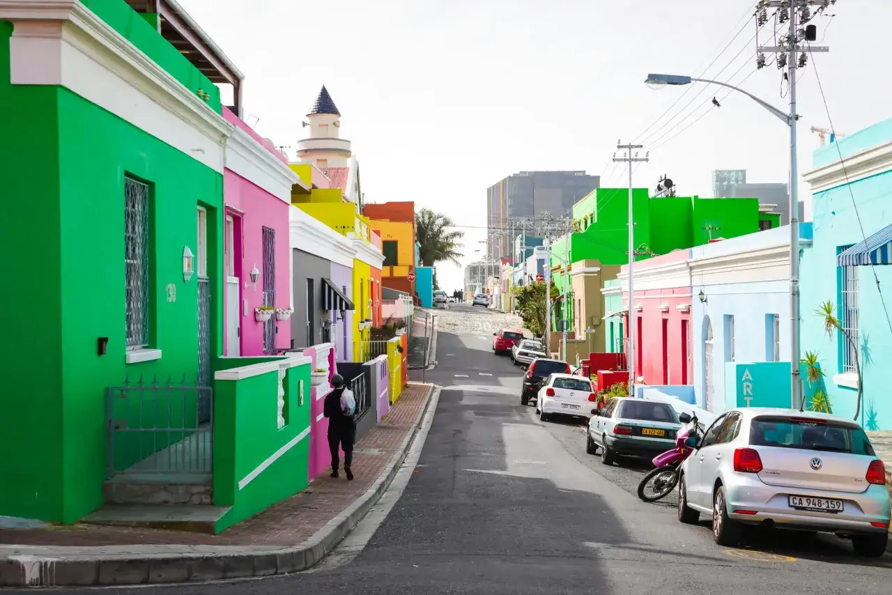 Rue colorée en Afrique du Sud, avec des maisons vives et des voitures garées. Un homme marche sur le trottoir, un moyen de transport typique de la région.
