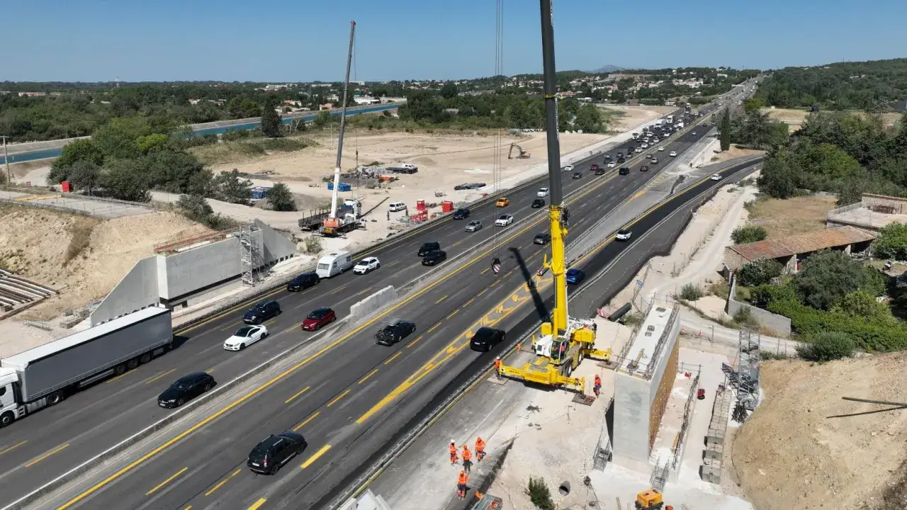 Chantier de création autoroute avec grues, camions et voitures circulant sur une voie en construction.