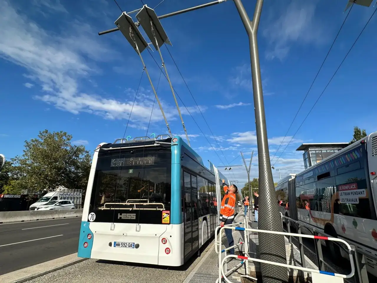 Un homme en gilet orange ajuste les perches d'un trolleybus bleu et blanc, connect&eacute; aux fils &eacute;lectriques a&eacute;riens.