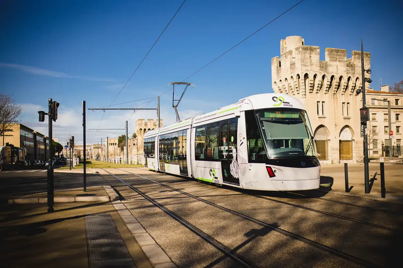 Le tramway moderne circule sur les rails à Avignon, passant devant des bâtiments historiques sous un ciel bleu éclatant.