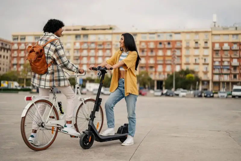 Deux jeunes femmes discutent, l'une sur un vélo blanc, l'autre sur une trottinette électrique. Elles profitent d'un moyen de transport doux en ville.