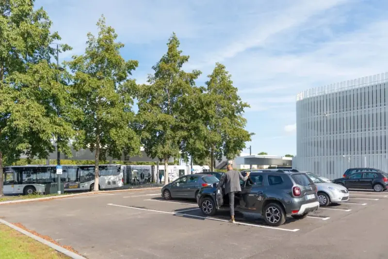 Un homme en costume monte dans un SUV gris dans un parking. Des bus électriques sont garés à proximité, près d'un bâtiment moderne. C'est une scène typique de naolib Nantes.