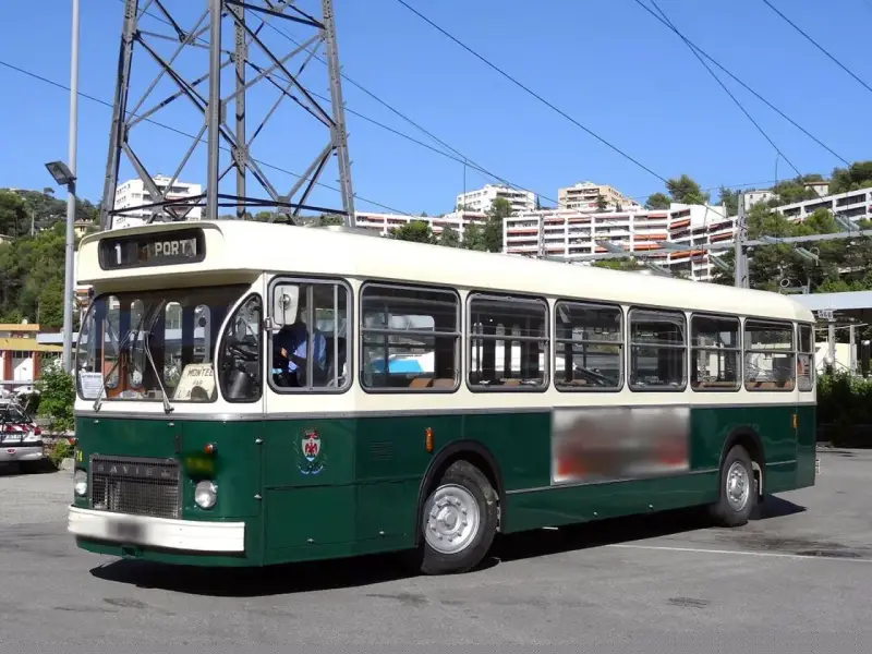 Un bus Saviem SC10, vert et crème, circule sous un ciel bleu. Il est stationné sur un parking, avec des immeubles en arrière-plan.